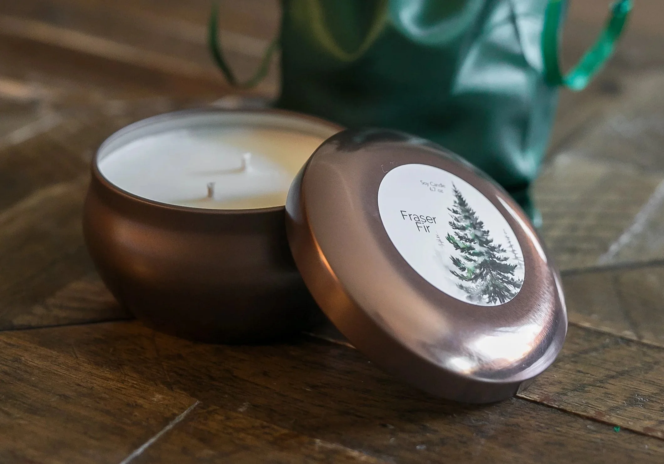 Open bronze-colored Fraser Fir candle tin with two wicks, its labeled lid leaning beside it and a green gift bag behind it.