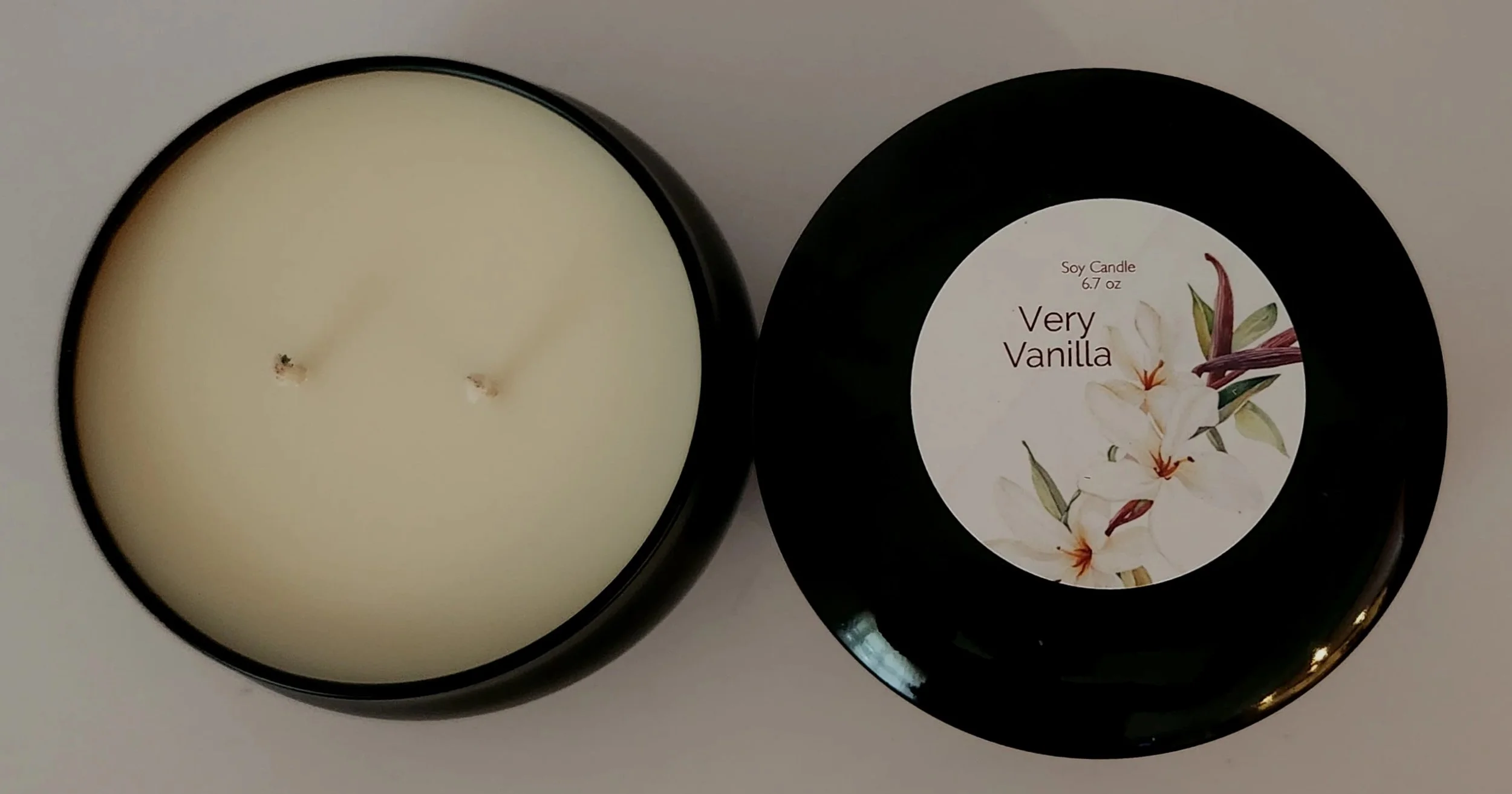 Overhead view of an open black Very Vanilla candle tin with two wicks beside its floral-labeled lid.