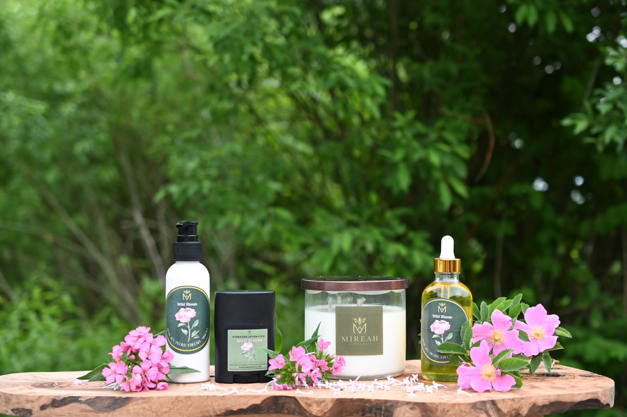 Mireah Wild Bloom bottles, a lotion stick and a candle arranged with pink flowers on a wooden board.