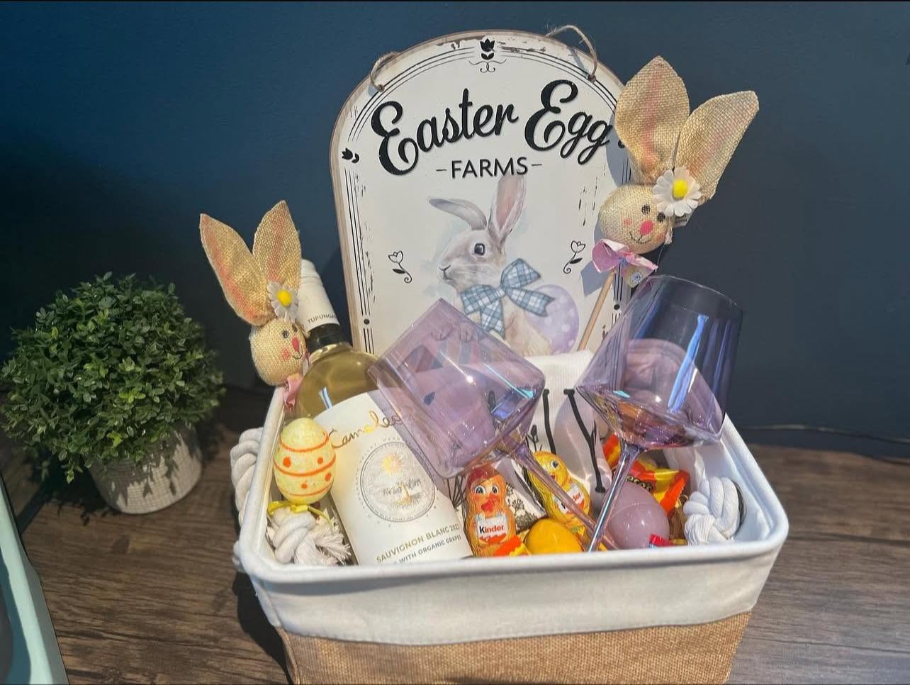 Fabric-lined Easter gift basket holding rabbit decorations, glasses and treats, with a decorative Easter sign behind them.